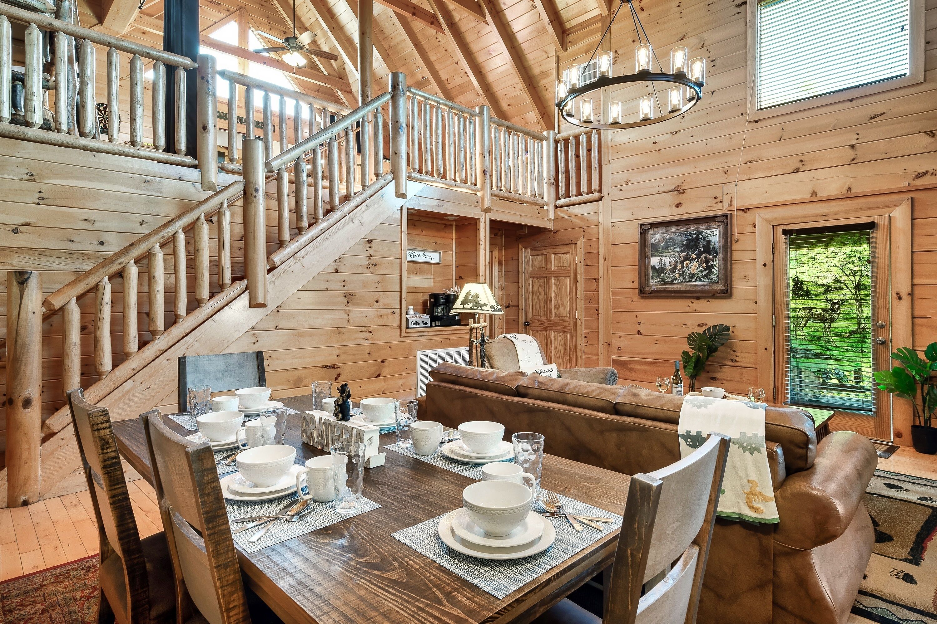 A wooden cabin interior with a dining area, a staircase, a sofa, and a window with a view of greenery.