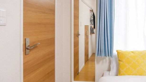 Quarto de hotel com porta de madeira, corredor com espelho e cama com travesseiro amarelo.