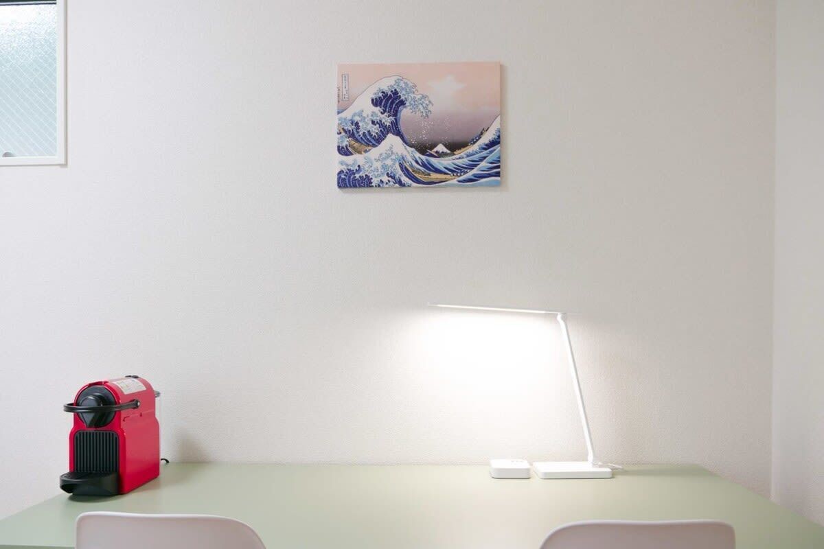 A room with a desk, a red device, a white desk lamp, and a Japanese wave painting on the wall.