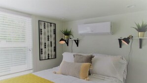 A bedroom with a bed, a bedside table, a wall-mounted air conditioner, and a window with blinds.