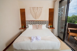 Habitación de hotel con una cama grande, un tapiz de macramé y vistas a la vegetación a través de un ventanal corredero.