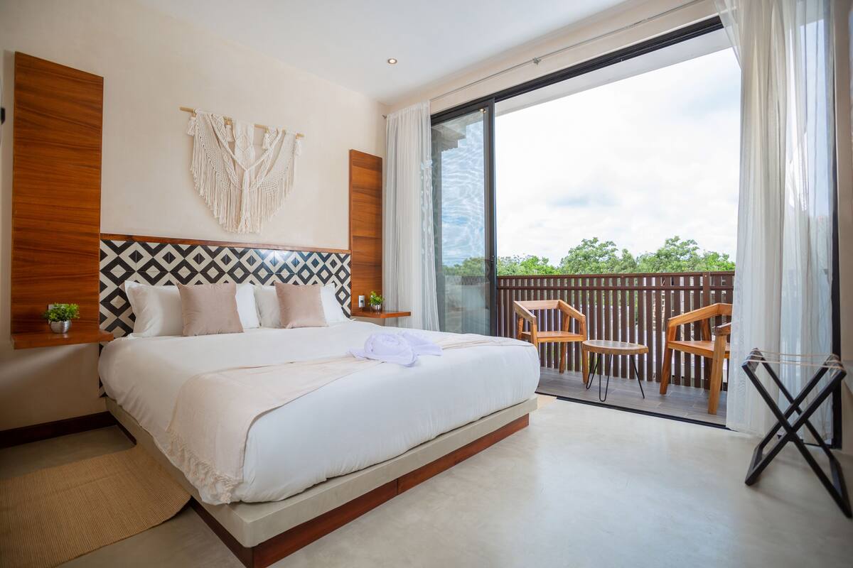 Um quarto moderno com uma cama grande, cabeceira de madeira, um painel de parede em macramé e uma varanda com cadeiras e mesa de madeira.