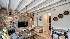 A modern living room with a brick wall, a flat-screen TV, a sofa, a coffee table, and a dining area with a wooden table and chairs.
