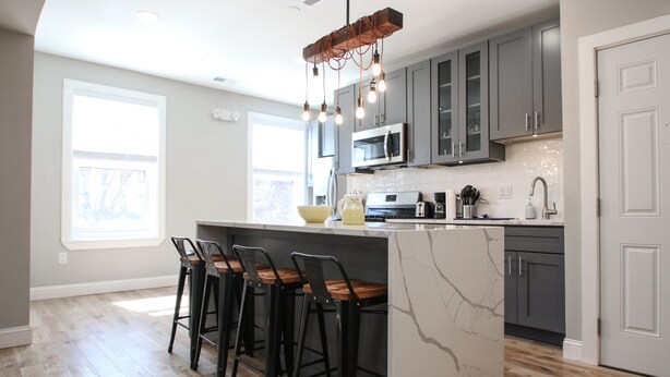 A modern kitchen with a marble island, bar stools, and a hanging light fixture.