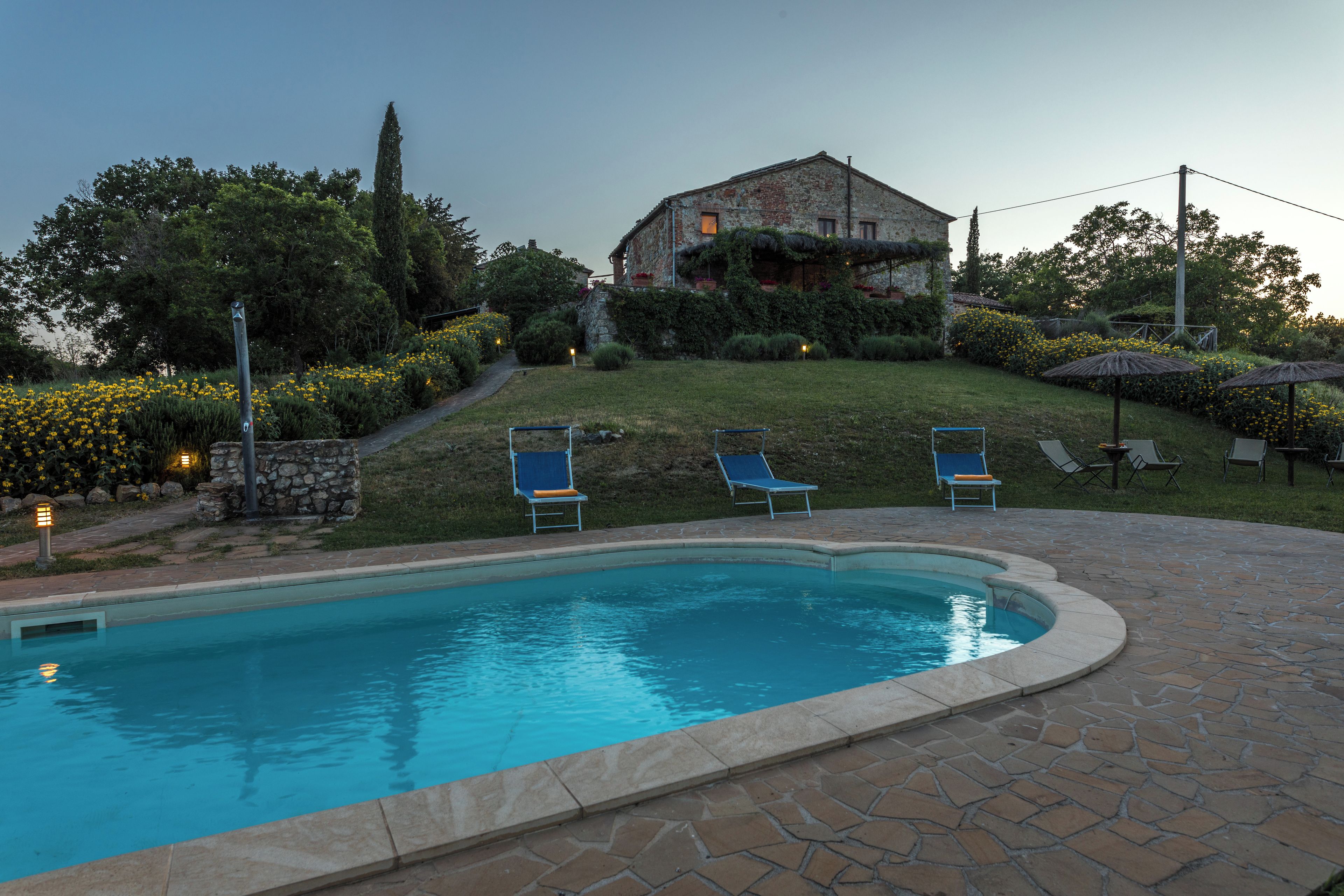 Una piscina con lettini, un edificio in pietra e un giardino con fiori gialli.