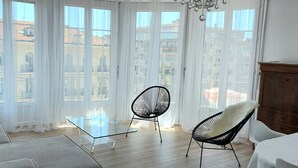 A modern room with large windows, a glass coffee table, two chairs, a wooden cabinet, and a view of buildings. - Le Métropole (Nice)