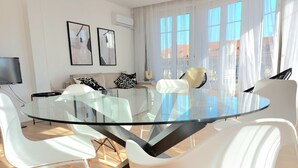 A modern living room with a glass-top dining table surrounded by white chairs, a sofa with patterned cushions, and a flat-screen TV. - Le Métropole (Nice)