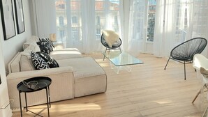 A modern living room with a sofa, two chairs, a glass coffee table, and large windows with a view of buildings. - Le Métropole (Nice)