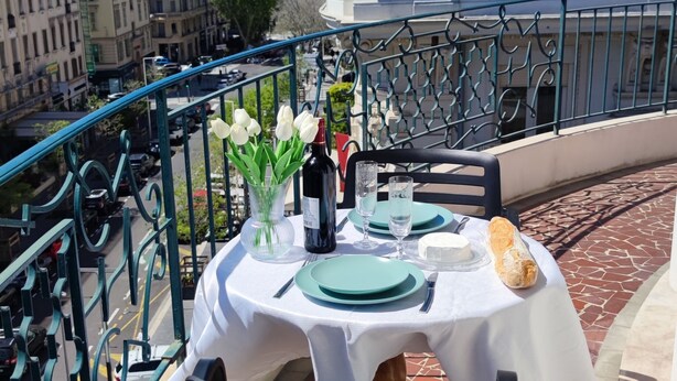 A table set with plates, cutlery, a bottle, and a vase of flowers on a balcony with a view of buildings. - Le Métropole (Nice)