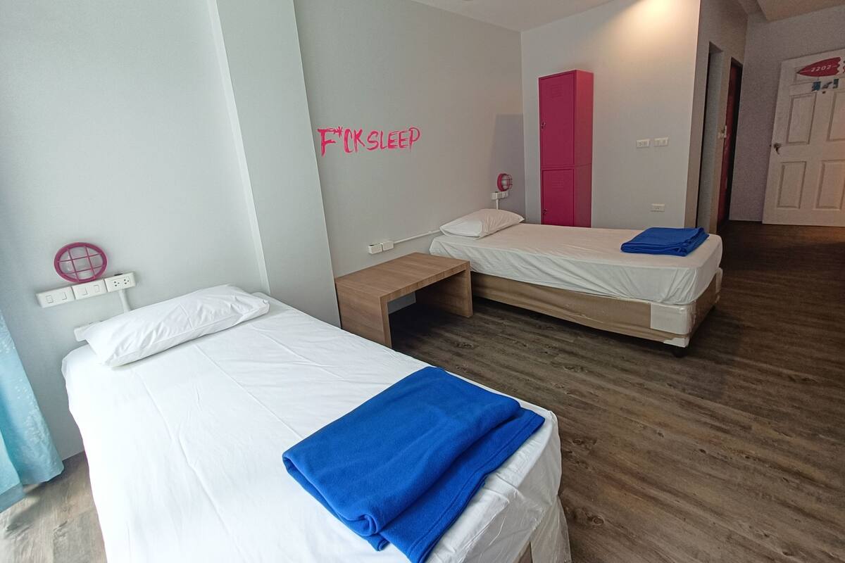 A hotel room with two single beds, a small wooden bench, a pink wardrobe, and a wall-mounted power outlet.