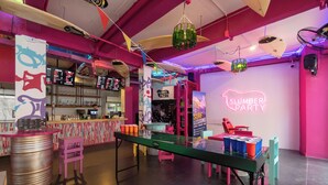 Een levendige bar met een neonbord waarop 'SLUMBER PARTY' staat, een lange toonbank met krukken en een neonbord voor 'SURF LESSONS'.