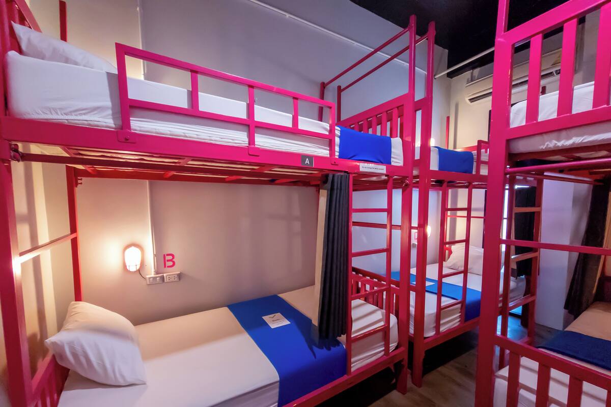 A bunk bed room with two beds, each with a pillow and a blue blanket. There is a wall socket and a light switch visible.