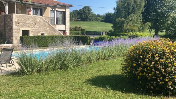 Una casa de piedra con piscina, rodeada de césped cuidado y arbustos en flor.