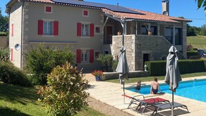 Una casa de dos plantas con piscina, contraventanas rojas y fachada de piedra.