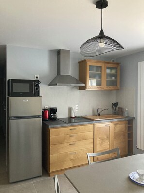 A compact kitchen with a built-in microwave, a stainless steel refrigerator, wooden cabinets, a sink, and a dining table with a chair. - Small single-storey apartment with PRM access, 2*. (Caylus)