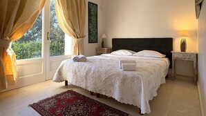 Une chambre à coucher avec un lit, des rideaux, une table de chevet, une lampe, un tapis et une fenêtre donnant sur un paysage verdoyant.