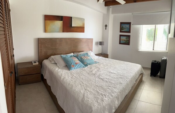Ein Schlafzimmer mit einem großen Bett, einem Holz-Kopfteil, einem Nachttisch mit Lampe, einem an der Wand befestigten Kreuz und einem Fenster mit Jalousien.