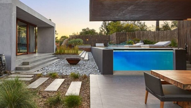 Un área de piscina moderna al aire libre con un deck de concreto, una mesa de madera y una silla de descanso.