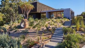 Una casa moderna con una escalera de piedra que lleva a la entrada, rodeada de vegetación nativa y árboles.