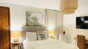 Een slaapkamer met een bed, twee nachtlampjes, een televisie, een rieten stoel en een schilderij aan de muur.