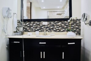 Un baño con salpicadero de azulejos blanco y negro, un lavabo blanco y un espejo encima. Hay un secador de pelo en la pared a la izquierda y una planta en la encimera.