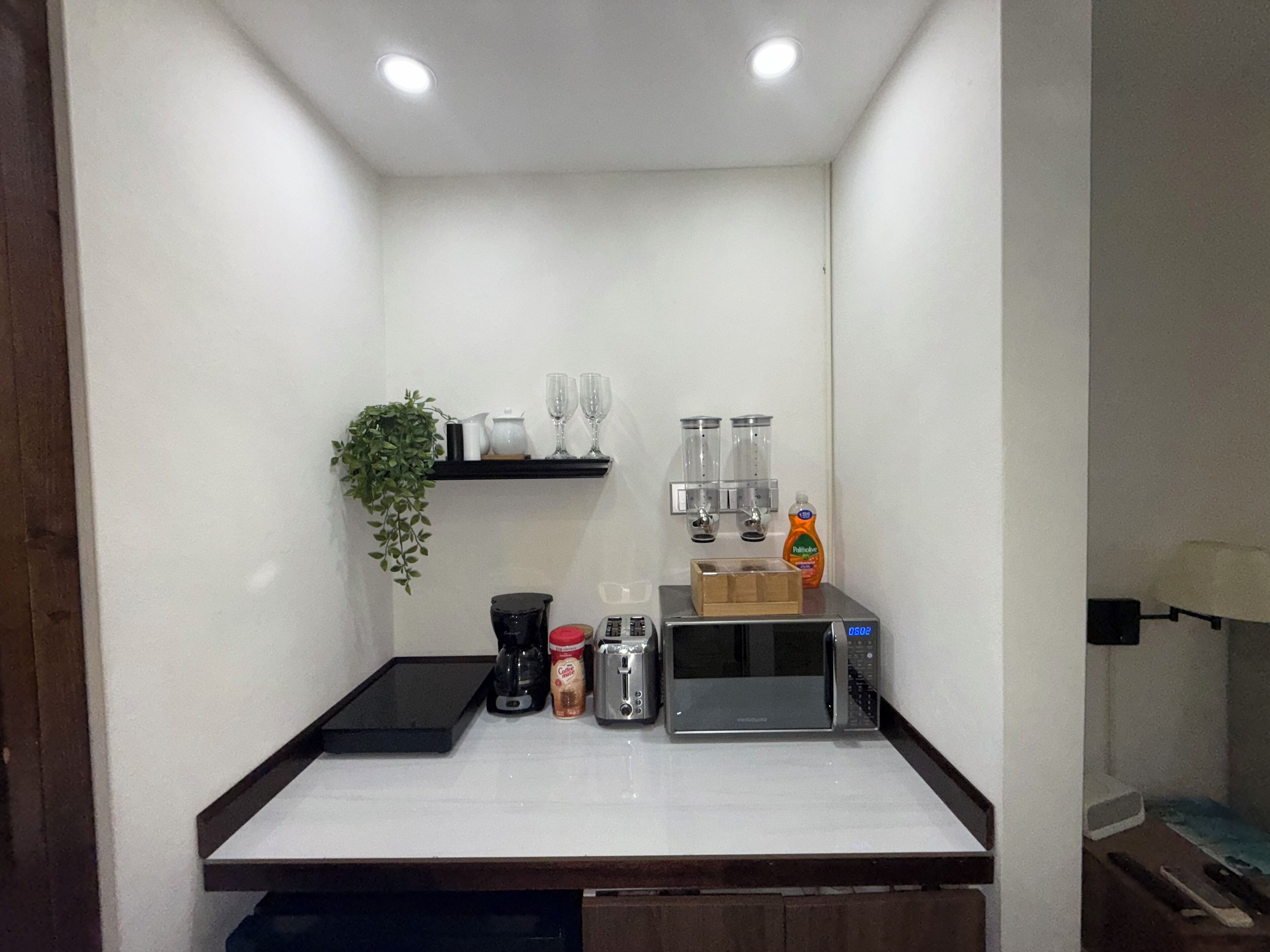 A compact kitchen with a microwave, toaster, coffee maker, and a shelf with glassware and a plant.