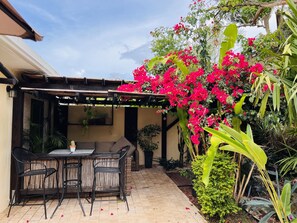 Un patio al aire libre con una mesa y sillas, rodeado de exuberante follaje y flores rosas vibrantes.