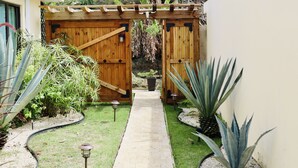 Ein gepflasterter Weg führt zu einem hölzernen Tor in einem Garten mit verschiedenen Pflanzen und zwei Laternen.