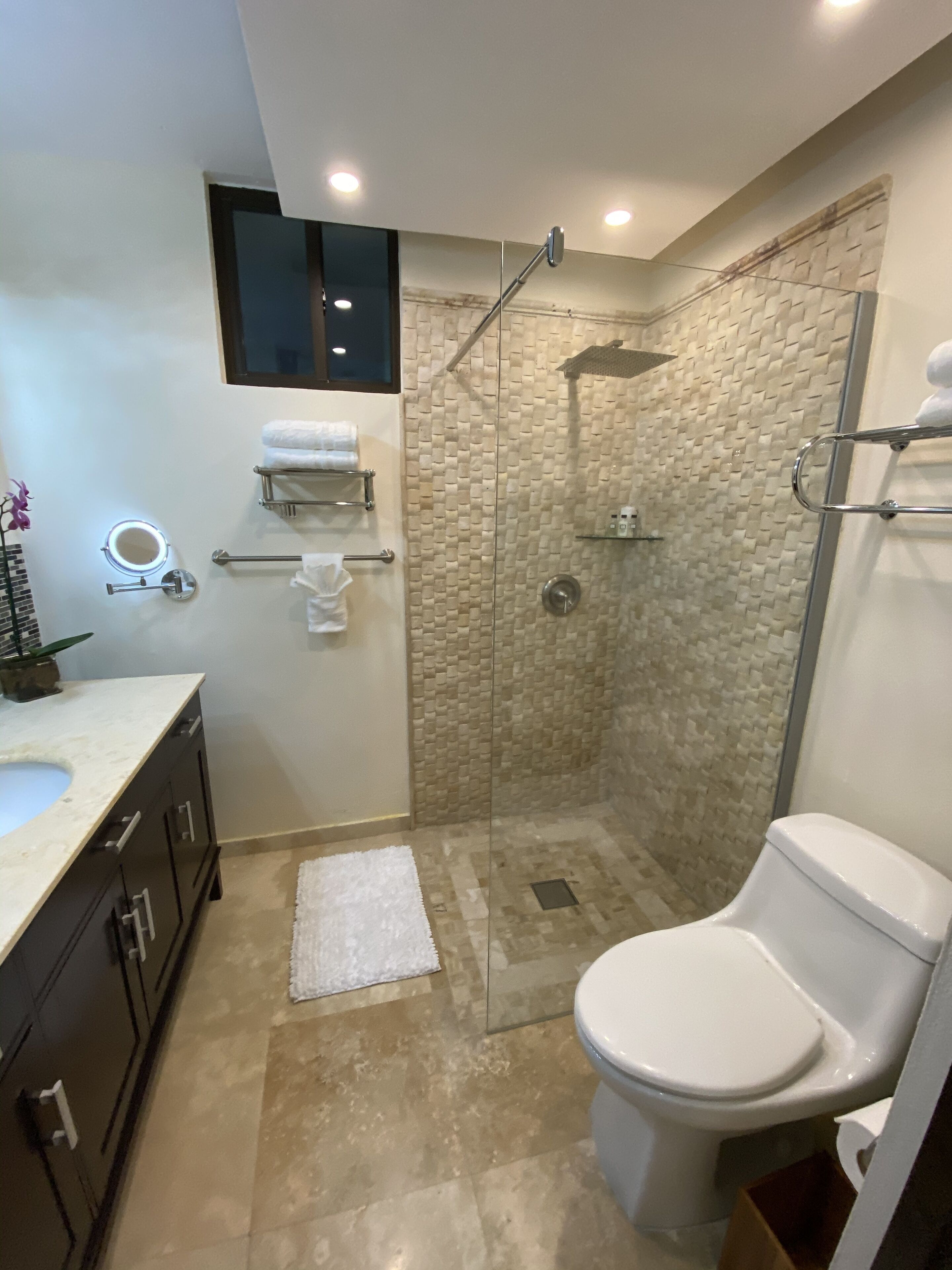 Une salle de bain moderne dotée d’une douche à parois de verre, de toilettes, d’un lavabo avec un plan de travail en marbre et d’un miroir avec éclairage intégré.