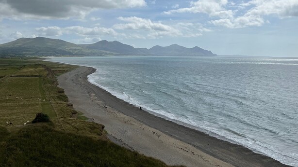 A coastal landscape with a beach, grassy fields, and distant mountains under a partly cloudy sky. - Awel-y-mor (Caernarfon)