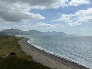 A coastal landscape with a beach, grassy fields, and distant mountains under a partly cloudy sky. - Awel-y-mor (Caernarfon)