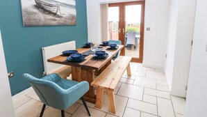A dining area with a wooden table set for four, a blue chair, and a bench. A painting of a boat is on the teal wall, and there's a view through a glass door to the outside. - Awel-y-mor (Caernarfon)
