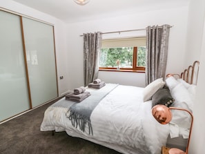 A bedroom with a bed, a window with curtains, a bedside table, and a view of greenery outside. - Awel-y-mor (Caernarfon)