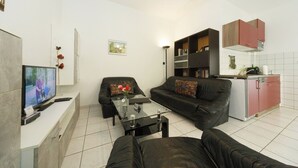 Ein modernes Wohnzimmer mit Sofa, Couchtisch und Fernseher. Es gibt eine Küchenzeile mit Schränken und einem Geschirrspüler.