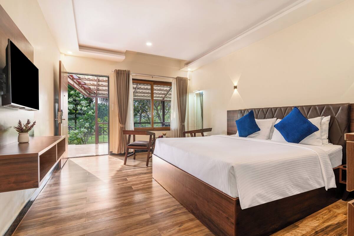 A modern hotel room with a large bed, wooden flooring, a TV, a desk, and a view of greenery.