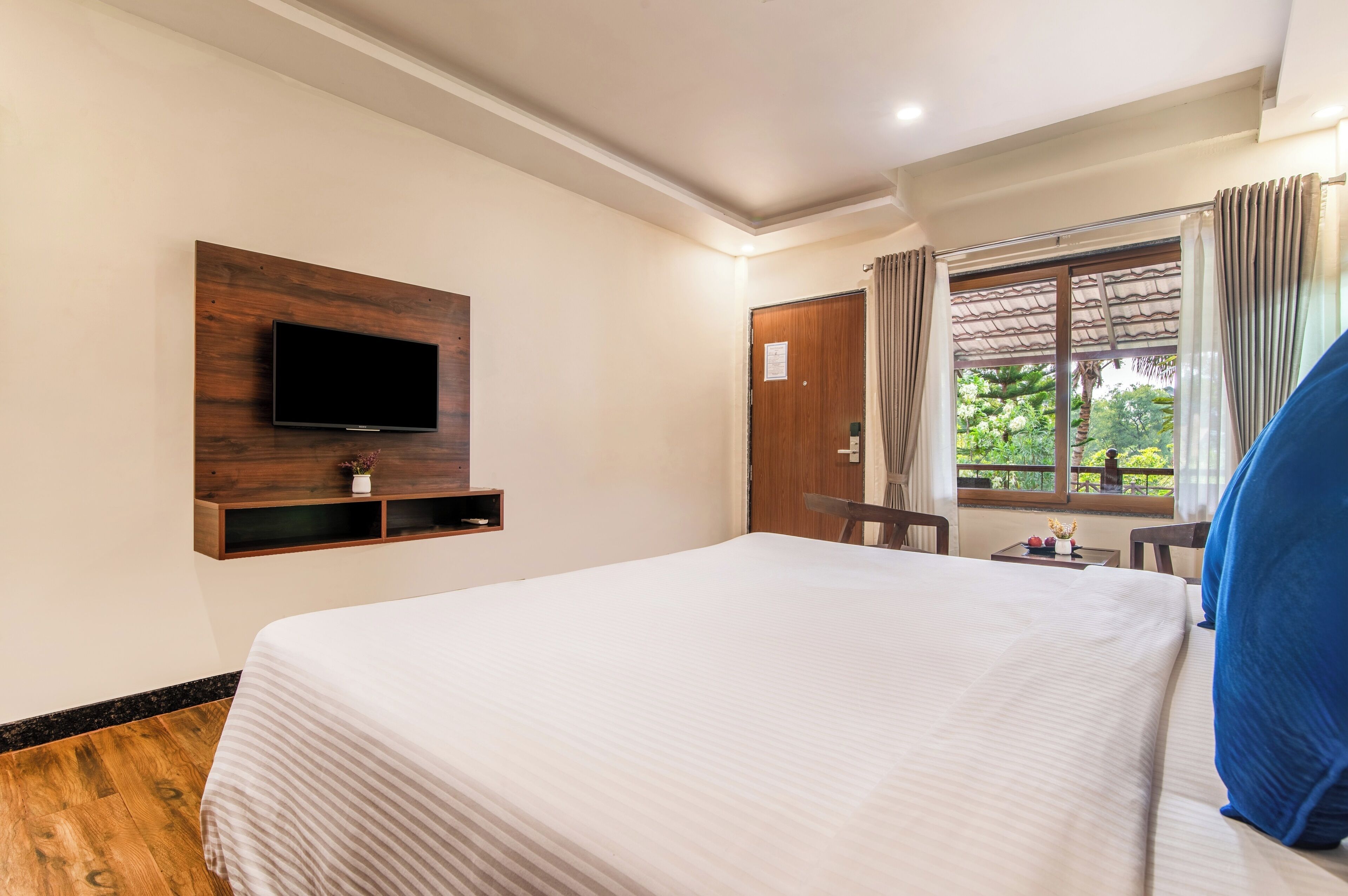 A hotel room with a bed, a TV mounted on a wooden panel, a window with curtains, and a small table with a vase.