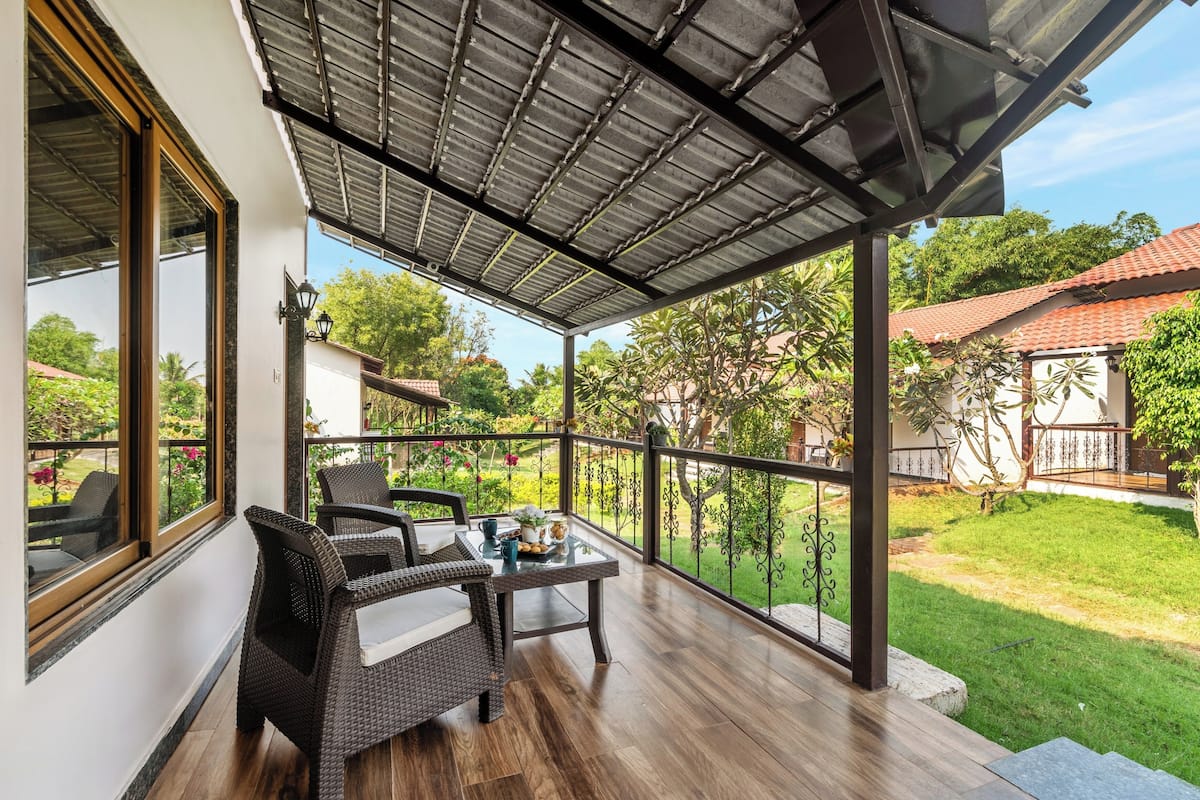 A covered outdoor area with wicker furniture and a table, overlooking a garden and neighboring houses.