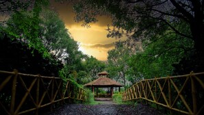 A rustic wooden gazebo with a thatched roof, surrounded by dense foliage and a bamboo fence. - Sterling Banashree Badami  (Badami)