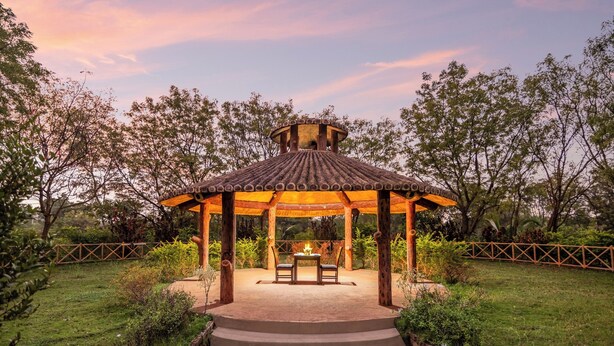 A gazebo with a tiled roof and a central table with chairs, surrounded by a garden with a wooden fence. - Sterling Banashree Badami  (Badami)