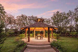 A gazebo with a tiled roof and a central table with chairs, surrounded by a garden with a wooden fence. - Sterling Banashree Badami  (Badami)