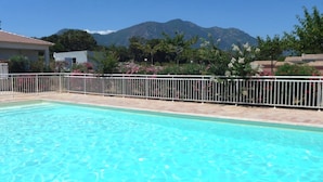 A swimming pool with a white fence, surrounded by a garden with pink flowers and a mountain in the background.