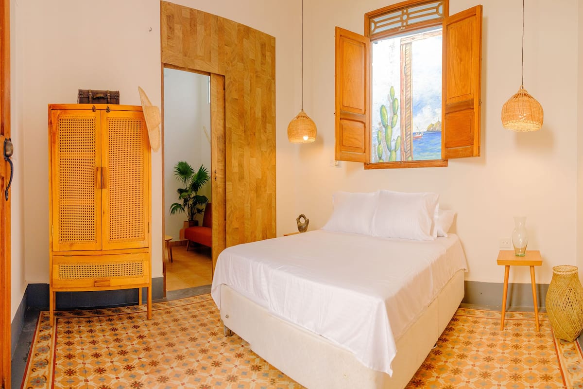 A bedroom with a bed, a wooden wardrobe, a window with open shutters, and a hanging light fixture.