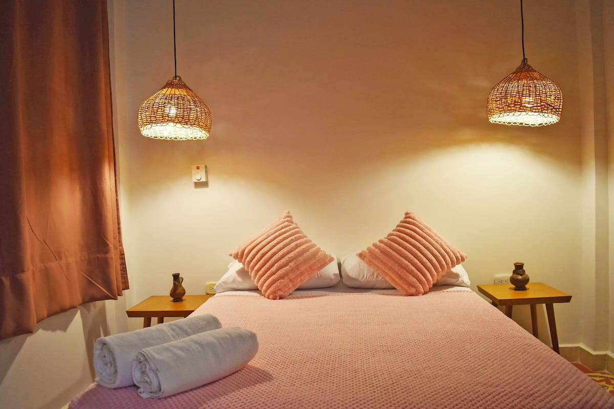 A neatly made bed with pink bedding, two bedside tables with decorative items, and two hanging lamps.