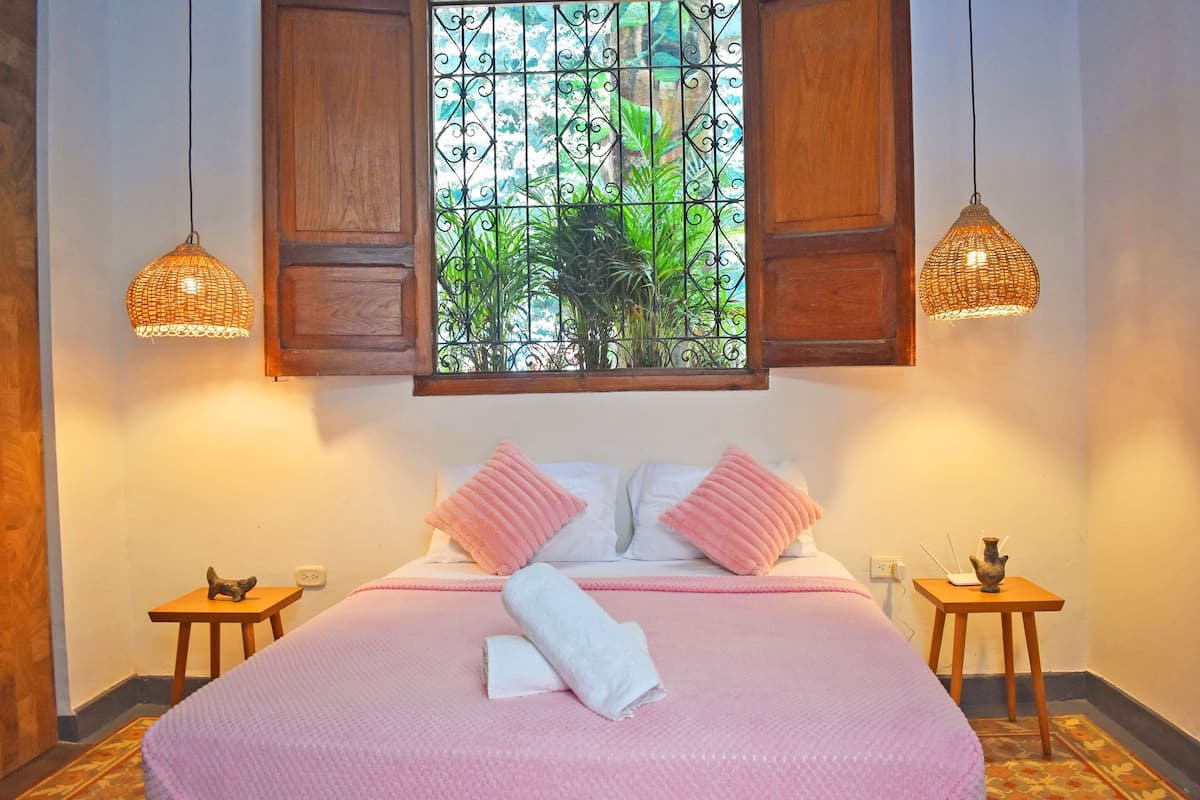A bedroom with a bed, two bedside tables, a window with a view of greenery, and two hanging lamps.