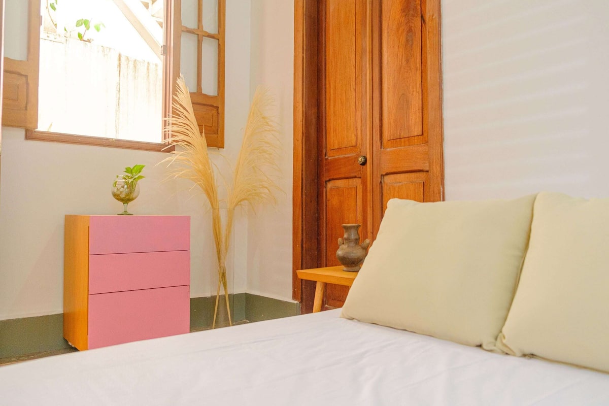 A bedroom with a bed, a wooden door, a pink and orange dresser, and a window with plants.
