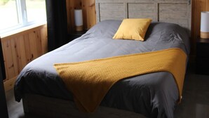 Une chambre à coucher avec une tête de lit en bois, un lit avec une literie grise et jaune, une fenêtre avec un rideau noir et une table de chevet avec une lampe.