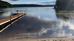 Une jetée en bois s’étendant dans une eau calme, entourée d’une plage de sable et d’une colline boisée.