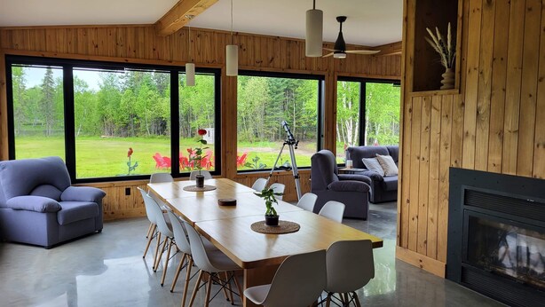 Un intérieur moderne avec un mur lambrissé en bois, de grandes fenêtres offrant une vue sur un paysage verdoyant, un coin repas avec une table et des chaises, et une cheminée.