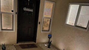 L'ingresso di una casa con una porta scura, tre finestre con persiane, un vialetto in cemento e piccoli vasi con piante.