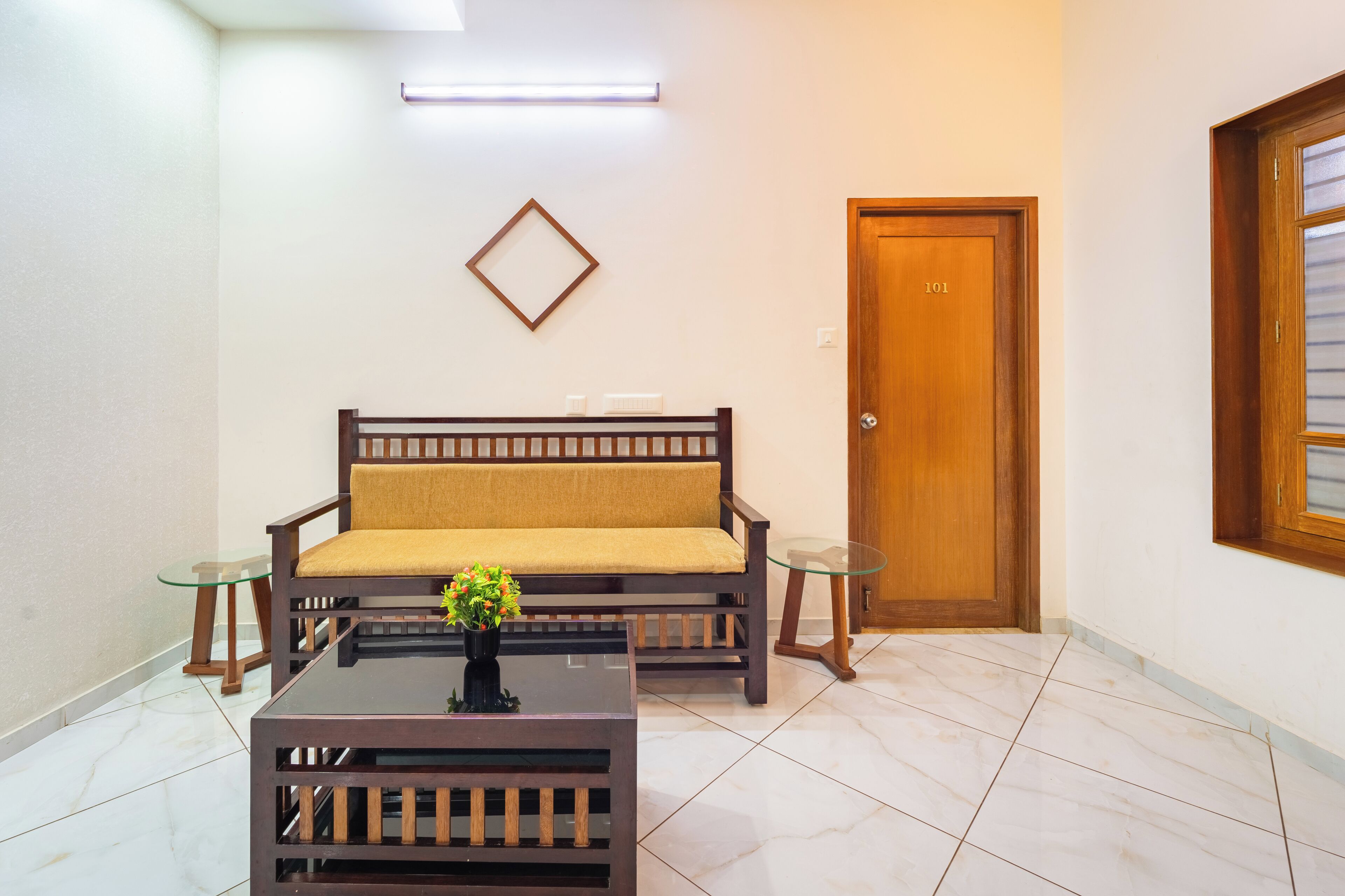 A room with a wooden sofa, a glass coffee table with a flower vase, and a wooden door with the number 101.